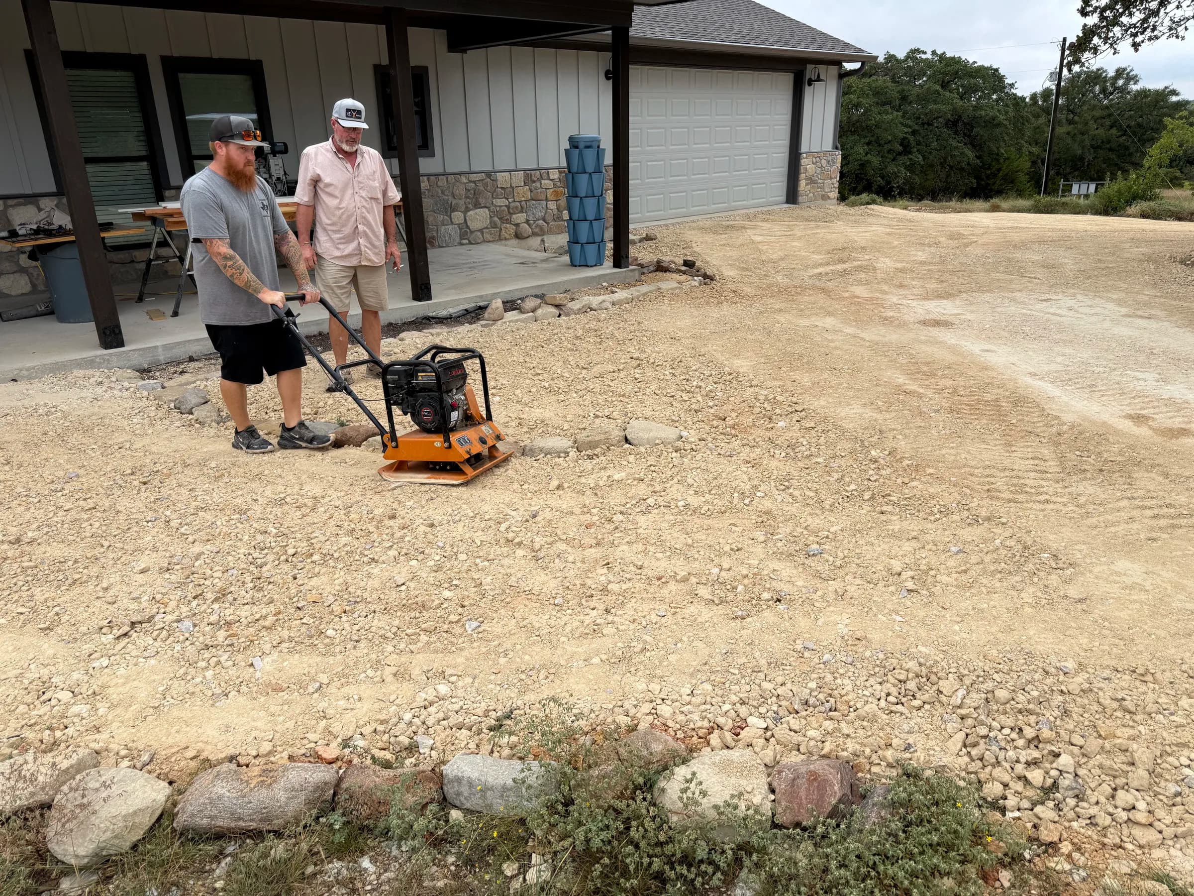 Two crew members working with a plate compactor beside the home in Ding Dong